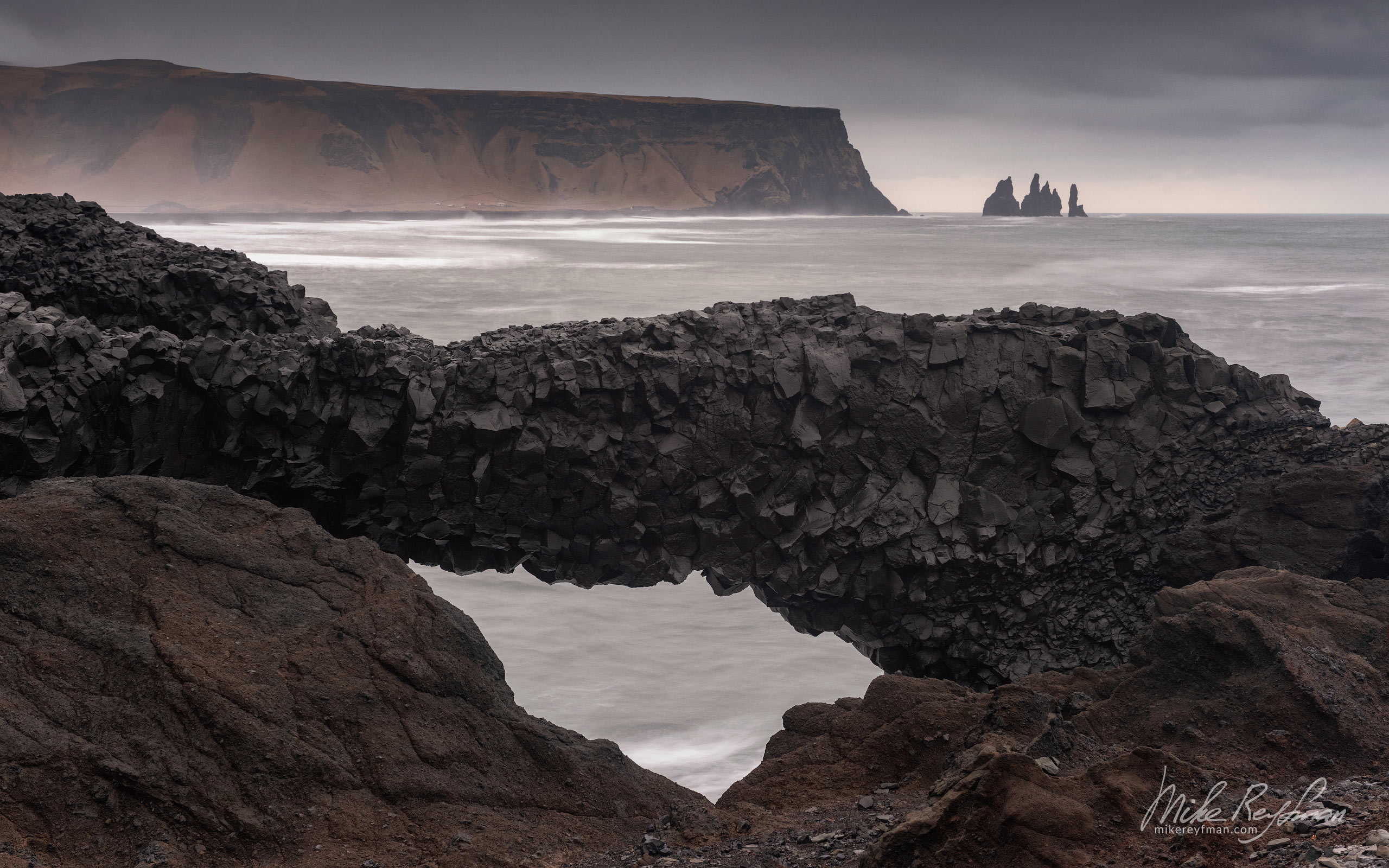 Distant view of Mt. Reynisfjall and Reynisdrangar basalt sea stacks from Cape Dyrholaey with basalt arch on the foreground. Southern Iceland. 024-IC-CL_10P2540 - Where Lava Meets the Ocean. Iceland coastline. - Mike Reyfman Photography
